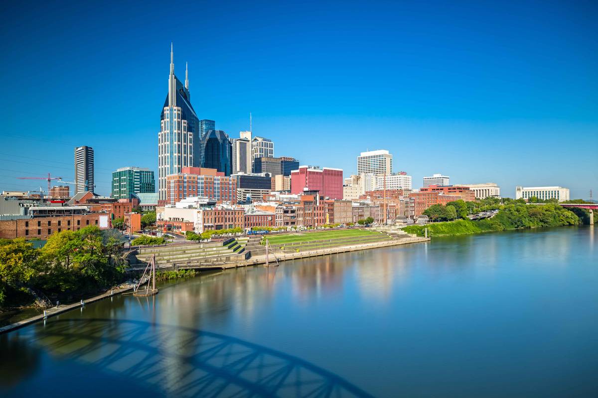 The Nashville skyline under a clear blue sky, seen across the Cumberland River, with the city's distinctive twin-spired tower rising at the left.
